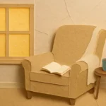 A cozy beige armchair by a sunlit window with a blanket draped over the arm and an open book resting on the seat, suggesting interrupted rest.